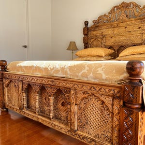 May include: A wooden bed frame featuring intricate hand-carved geometric and floral patterns on the headboard and footboard, paired with a light gold patterned bedspread and matching pillows, set against a plain white wall.