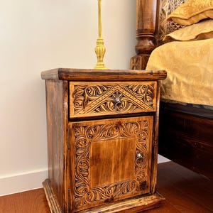 May include: A dark brown wooden nightstand featuring intricate hand-carved floral patterns on the drawer front and the cabinet door. The piece includes a single drawer with a metal pull and a lower cabinet door with a matching metal pull.