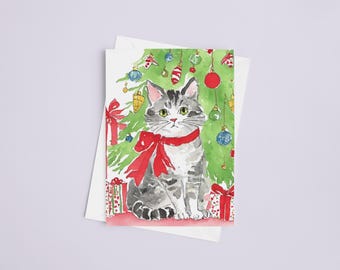 Pode incluir: Cartão de Natal em aquarela com um gato cinza tigrado com um laço vermelho, sentado em frente a uma árvore de Natal decorada. Os presentes estão na base da árvore. Enfeites vermelhos, azuis e dourados pendurados na árvore.