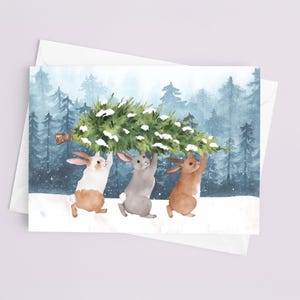 May include: Watercolor illustration of three rabbits carrying a Christmas tree through a snowy forest. The rabbits are in shades of white, gray, and brown. The background features a blue and green forest scene, and the image is on a white card.