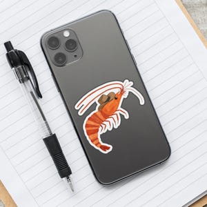 May include: A gray iPhone with a sticker of a shrimp wearing a cowboy hat. The shrimp is orange and has a white shell. The sticker is on the back of the phone.