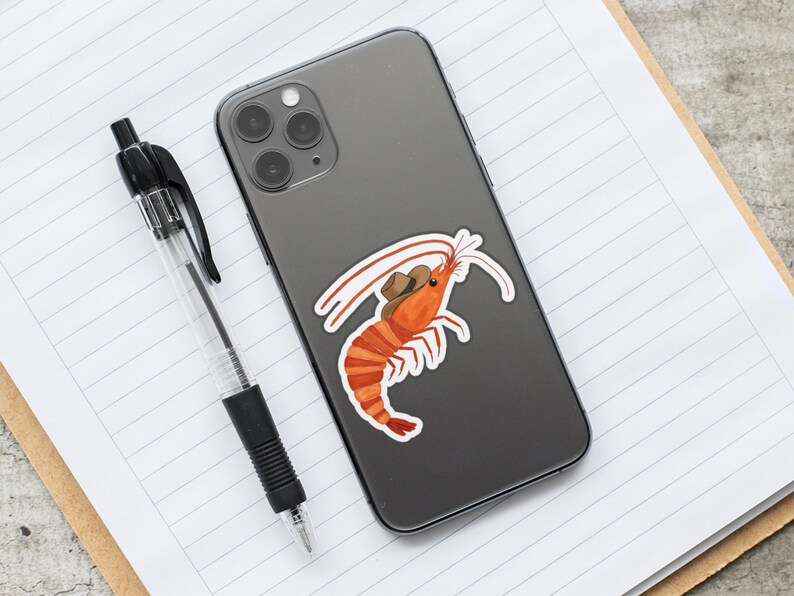 May include: A gray iPhone with a sticker of a shrimp wearing a cowboy hat. The shrimp is orange and has a white shell. The sticker is on the back of the phone.