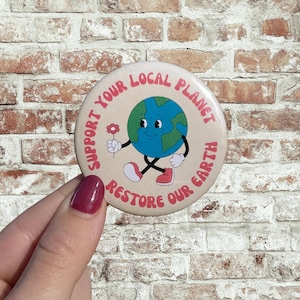 Könnte beinhalten: Ein runder Button mit dem Text "Support Your Local Planet, Restore Our Earth". Der Button zeigt einen Cartoon-Erdcharakter, der eine Blume hält. Die Erde ist blau und grün, mit einem lächelnden Gesicht. Der Button wird in einer Hand gehalten.