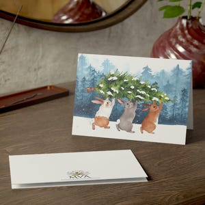 Pode incluir: Um cart&atilde;o de Natal com uma ilustra&ccedil;&atilde;o em aquarela de tr&ecirc;s coelhos carregando uma &aacute;rvore de Natal por uma floresta nevada. Outro cart&atilde;o &eacute; vis&iacute;vel em primeiro plano. Um vaso e um espelho est&atilde;o no fundo.