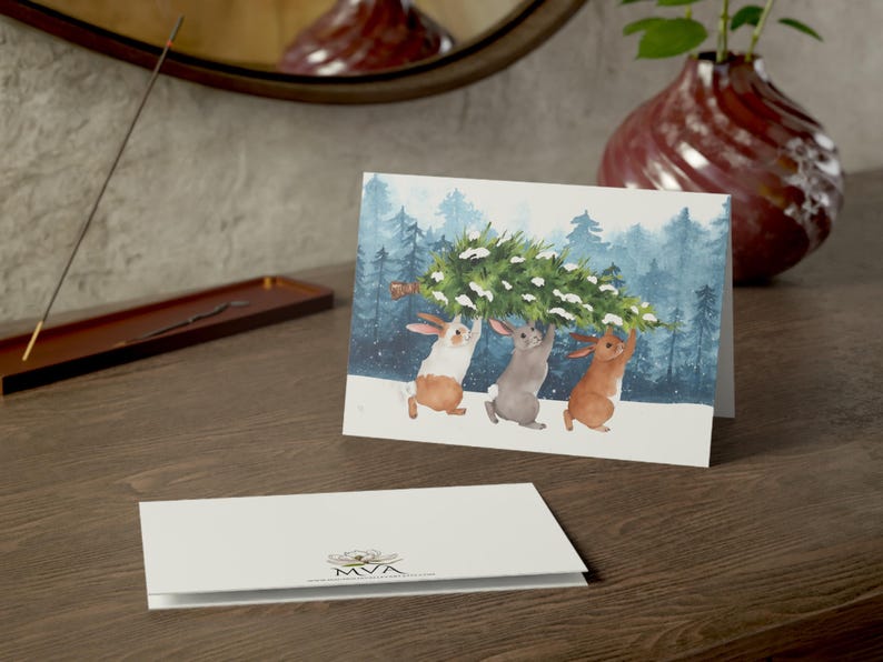 Pode incluir: Um cart&atilde;o de Natal com uma ilustra&ccedil;&atilde;o em aquarela de tr&ecirc;s coelhos carregando uma &aacute;rvore de Natal por uma floresta nevada. Outro cart&atilde;o &eacute; vis&iacute;vel em primeiro plano. Um vaso e um espelho est&atilde;o no fundo.