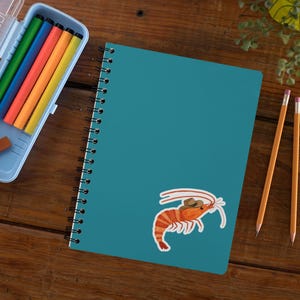 May include: A teal blue spiral notebook with a sticker of a cartoon shrimp wearing a cowboy hat. The notebook is on a wooden surface with two yellow pencils.