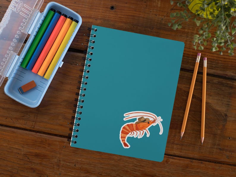May include: A teal blue spiral notebook with a sticker of a cartoon shrimp wearing a cowboy hat. The notebook is on a wooden surface with two yellow pencils.