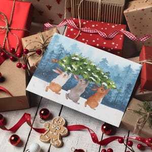 Pode incluir: Uma imagem com tema natalino com uma ilustra&ccedil;&atilde;o em aquarela de tr&ecirc;s coelhos carregando uma &aacute;rvore de Natal. Rodeada por presentes embrulhados, fitas vermelhas e enfeites, criando uma cena festiva de feriado.