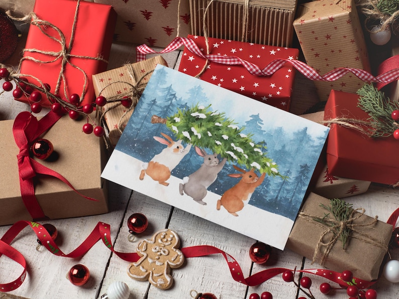 Pode incluir: Uma imagem com tema natalino com uma ilustra&ccedil;&atilde;o em aquarela de tr&ecirc;s coelhos carregando uma &aacute;rvore de Natal. Rodeada por presentes embrulhados, fitas vermelhas e enfeites, criando uma cena festiva de feriado.
