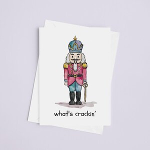 May include: A watercolour illustration of a nutcracker soldier on a white card. The nutcracker wears a pink jacket, blue trousers, and a crown. The text "what's crackin'" is below the illustration. The card is on a white background.
