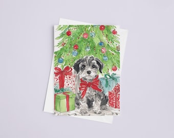 Pode incluir: Cartão de Natal em aquarela com um cão cinza e branco com um laço vermelho, sentado sob uma árvore de Natal decorada. Os presentes embrulhados em vermelho, verde e azul estão na base da árvore. O cartão está em um fundo branco.