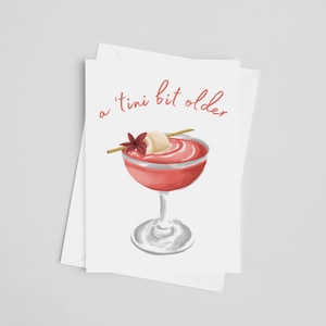 Può includere: Un biglietto bianco con il testo "a 'tini bit older" in lettere rosa. Un cocktail rosa con una guarnizione bianca è illustrato sulla carta.