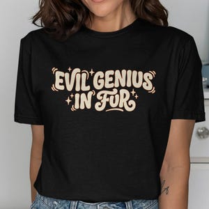 Puede incluir: Camiseta negra con el texto "EVIL GENIUS IN FUR" en crema con una fuente retro. El texto está delineado en un color más oscuro. La camiseta tiene cuello redondo y mangas cortas.
