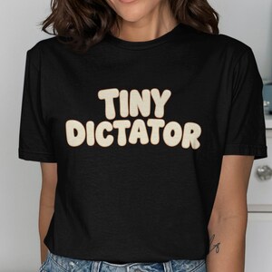 以下が含まれることがあります： レトロなバブルスタイルのフォントで「TINY DICTATOR」という文字がプリントされた黒いTシャツ。テキストは明るいベージュ色で、暗い背景と対照的です。カジュアルウェアアイテム。