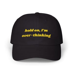 May include: Black baseball cap with the embroidered text "hold on, I'm over-thinking" in yellow. The cap has a curved brim and a structured fit, suitable for casual wear.