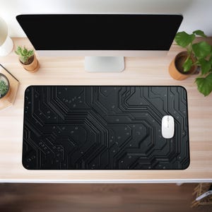 May include: Black rectangular mouse mat featuring a circuit board pattern. A white computer mouse is positioned on the mat. The mat sits on a light wood desk, with a computer monitor and plants in the background.