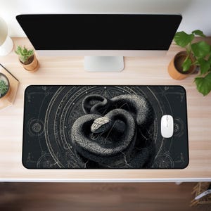 Puede incluir: Alfombrilla de ratón rectangular negra con una ilustración detallada de una serpiente enroscada con cabeza blanca. La serpiente está sobre un fondo oscuro con elementos de diseño geométricos y celestiales. Un ratón de ordenador blanco está al lado.