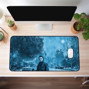 May include: A rectangular mousepad featuring a blue-toned image of a horror movie scene. The image includes a figure in a mask, cars, trees, and pumpkins. A white computer mouse is on the right side of the mousepad.