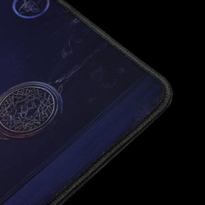 Puede incluir: Una alfombrilla de rat&oacute;n azul oscuro con un borde cosido negro. La alfombrilla presenta un dise&ntilde;o circular con un patr&oacute;n detallado y ornamentado en plata y oro. Un dise&ntilde;o circular m&aacute;s peque&ntilde;o es visible en la esquina superior izquierda.