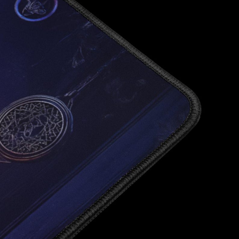 Puede incluir: Una alfombrilla de rat&oacute;n azul oscuro con un borde cosido negro. La alfombrilla presenta un dise&ntilde;o circular con un patr&oacute;n detallado y ornamentado en plata y oro. Un dise&ntilde;o circular m&aacute;s peque&ntilde;o es visible en la esquina superior izquierda.