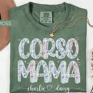 May include: A moss green t-shirt with the words "CORSO MAMA" in a floral design with pink bows. The shirt also features the names "charlie & daisy" in a script font. A gold star pendant necklace rests on the shirt.