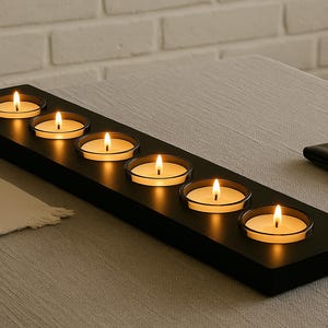 Może przedstawiać: Czarny, prostokątny świecznik z sześcioma zapalonymi świecami typu tealight. Świece są w przezroczystych szklanych kubkach, a płomienie są jasne. Świecznik stoi na szarej serwetce, w tle biała ceglana ściana.