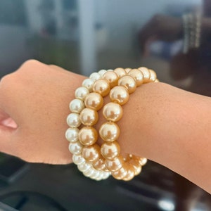 Op de afbeelding: Een set van twee gelaagde armbanden gemaakt van ronde, gladde kralen. De ene armband heeft glanzende witte kralen, terwijl de andere bestaat uit metallic champagnekleurige kralen. De kralen zijn uniform van grootte en in één rij gerangschikt.