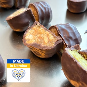 Op de afbeelding: Verschillende stukjes met pure chocolade omhulde lekkernijen met zichtbare vullingen van noten en granen, waarvan sommige doormidden zijn gesneden om een textuurrijke binnenkant te tonen. Een vierkant label toont de tekst MADE in Ukraine en een hart.