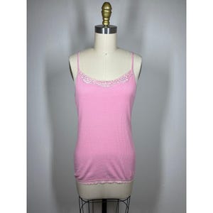 May include: A light pink camisole with thin spaghetti straps and delicate lace trim along the neckline and hem. The top is displayed on a mannequin, showcasing its simple, feminine design. The fabric appears to be a soft knit.