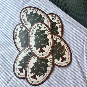 May include: A stack of oval stickers with a green tree and the text "Stick to your roots" in a brown circle with a white background. The stickers have a brown, orange, and white border.