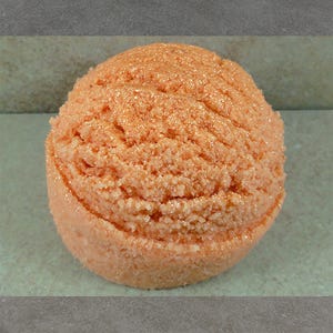 May include: A close-up of a round, peach-colored bath bomb with a textured surface and a sparkling, glittery appearance. The bath bomb is sitting on a light green surface.