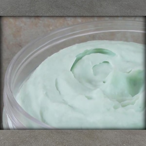 May include: Close-up of a clear plastic container filled with a light green, creamy substance. The cream has a smooth, whipped texture with visible swirls and a slight sheen. The container is partially visible.