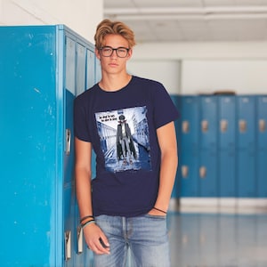 Pu&ograve; includere: Una t-shirt blu navy con una stampa grafica di una persona con un trench coat che cammina lungo un corridoio. Il testo sulla t-shirt recita "The devil is in the details".