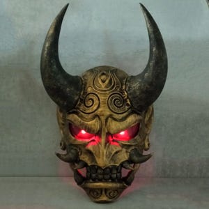 May include: A wooden Japanese demon mask with black horns and red glowing eyes. The mask has intricate carvings and a detailed design.