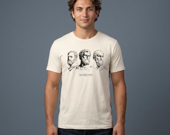 May include: A cream-colored short-sleeve t-shirt featuring a black graphic print of three bearded classical philosophers in profile. Below the illustrations, the word MORONS is printed in a clean, black, serif font centered on the chest area.