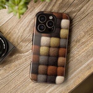May include: A smartphone case with a textured, quilted design in shades of brown, beige, and gray. The case has a glossy black border and a camera cutout. The phone case is resting on a wooden surface.