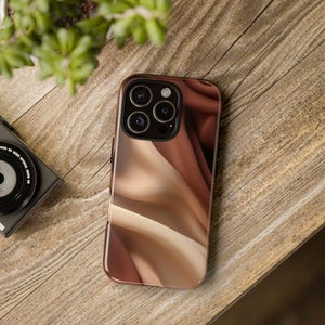 May include: A smartphone case with a flowing abstract design in shades of brown and beige. The case is on a wooden surface, with a camera and a plant in the background. The case has a glossy finish.