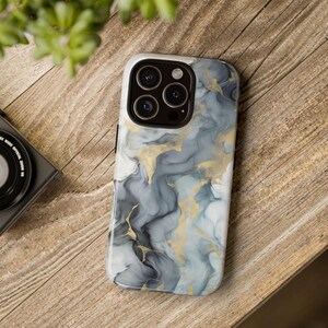 May include: A smartphone case with a marble design in shades of gray, white, and gold. The case has a glossy finish and a camera cutout. The phone case is on a wooden surface.