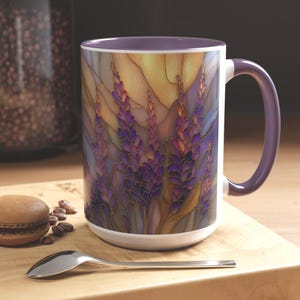May include: White ceramic mug with a purple interior and handle. The mug features a stained-glass style design of purple lavender flowers with gold accents against a warm-toned background. A spoon and a macaron are on a wooden surface.