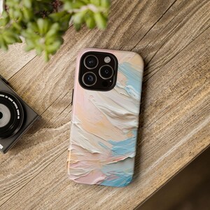 May include: A smartphone case with a pastel abstract art design in shades of pink, blue, and peach. The case is on a wooden surface, with a camera and plant in the background. The case has a glossy finish.