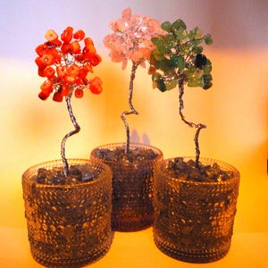May include: Three small potted trees made of wire and gemstones. The trees are in clear glass pots with a textured surface. The trees are decorated with red, pink, and green gemstones.