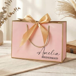 Puede incluir: Una bolsa de regalo rosa con lazo y asas doradas. La bolsa tiene el nombre "Amelia" y la palabra "Bridesmaid" impresa. La bolsa est&aacute; sobre una superficie beige.
