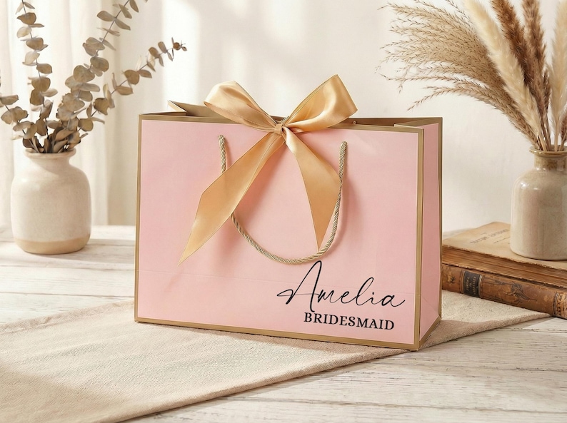 Puede incluir: Una bolsa de regalo rosa con lazo y asas doradas. La bolsa tiene el nombre "Amelia" y la palabra "Bridesmaid" impresa. La bolsa est&aacute; sobre una superficie beige.