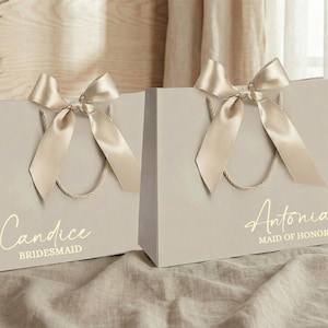 Puede incluir: Dos bolsas de regalo gris claro con lazos de sat&eacute;n dorado y asas. Una bolsa dice "Candice, Bridesmaid" y la otra "Antonia, Maid of Honor" en letras doradas. Las bolsas est&aacute;n sobre una superficie de tono neutro.