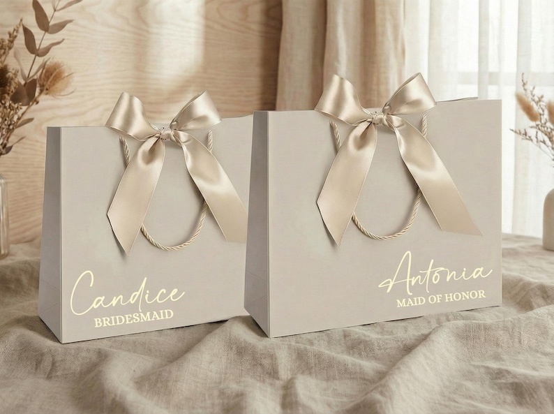 Puede incluir: Dos bolsas de regalo gris claro con lazos de sat&eacute;n dorado y asas. Una bolsa dice "Candice, Bridesmaid" y la otra "Antonia, Maid of Honor" en letras doradas. Las bolsas est&aacute;n sobre una superficie de tono neutro.
