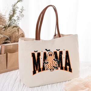 K&ouml;nnte beinhalten: Cremefarbene Tragetasche mit braunen Ledergriffen. Die Tasche zeigt das Wort "MAMA" in schwarzen und orangefarbenen Blockbuchstaben mit einer karierten Geistergrafik. Kleine schwarze Flederm&auml;use befinden sich &uuml;ber dem Wort.