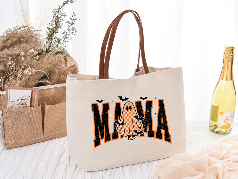 K&ouml;nnte beinhalten: Cremefarbene Tragetasche mit braunen Ledergriffen. Die Tasche zeigt das Wort "MAMA" in schwarzen und orangefarbenen Blockbuchstaben mit einer karierten Geistergrafik. Kleine schwarze Flederm&auml;use befinden sich &uuml;ber dem Wort.