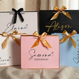 Puede incluir: Bolsas de regalo en blanco, negro, rosa y azul, cada una con un nombre en escritura elegante y un lazo de cinta a juego. Las bolsas son para damas de honor, con la palabra "Dama de honor" impresa debajo.