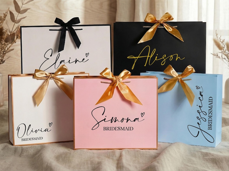 Puede incluir: Bolsas de regalo en blanco, negro, rosa y azul, cada una con un nombre en escritura elegante y un lazo de cinta a juego. Las bolsas son para damas de honor, con la palabra "Dama de honor" impresa debajo.
