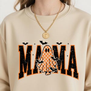 K&ouml;nnte beinhalten: Beiges Sweatshirt mit dem Wort "MAMA" in verwitterten schwarzen und orangefarbenen Buchstaben. Eine karierte Geistergrafik befindet sich in der Mitte, umgeben von Flederm&auml;usen. Das Sweatshirt ist ein Halloween-Design.